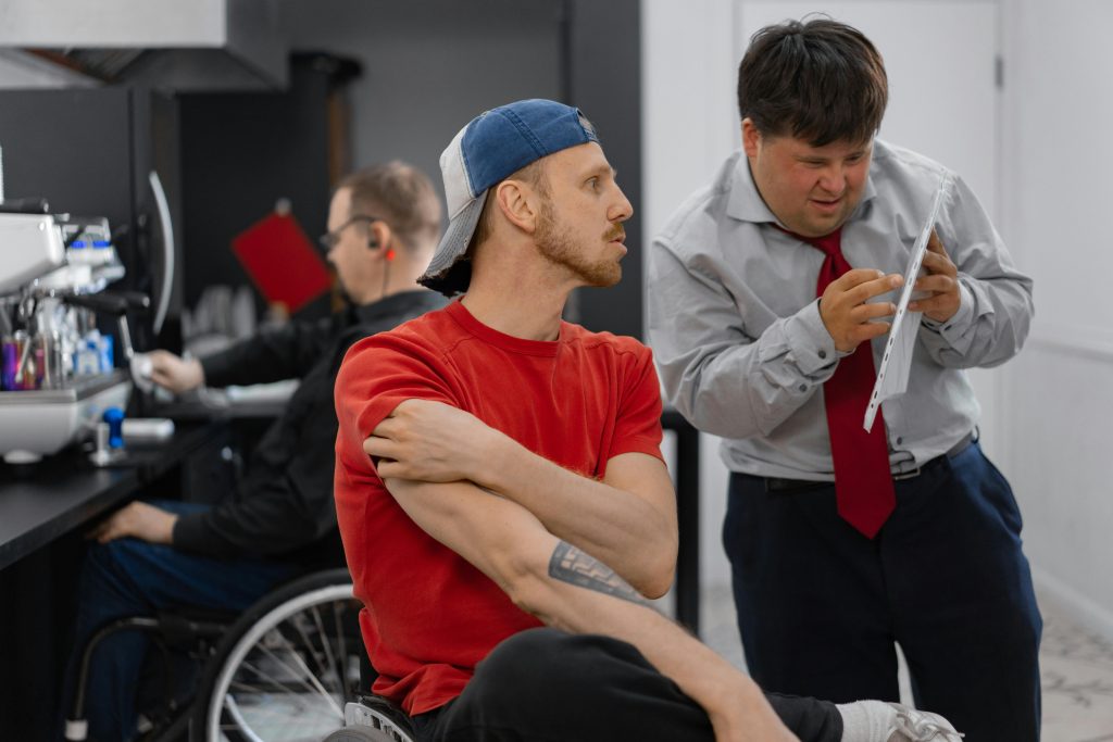 Twee mannen tijdens een workshop: een man in een stoel met rood t-shirt en blauwe pet kijkt geconcentreerd opzij, terwijl een man in grijs overhemd met rode stropdas achter hem staat en een vel papier bestudeert. Op de achtergrond zijn andere workshopdeelnemers bezig.