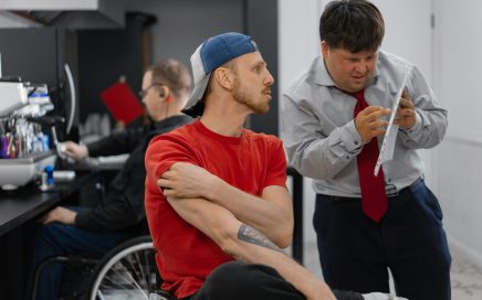 Twee mannen tijdens een workshop: een man in een stoel met rood t-shirt en blauwe pet kijkt geconcentreerd opzij, terwijl een man in grijs overhemd met rode stropdas achter hem staat en een vel papier bestudeert. Op de achtergrond zijn andere workshopdeelnemers bezig.